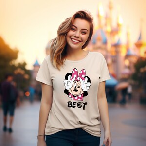 Może przedstawiać: Beżowa koszulka z nadrukiem kresk&oacute;wkowej postaci Minnie Mouse w r&oacute;żowej kokardce, kt&oacute;ra pokazuje znak pokoju dłońmi. Pod obrazkiem znajduje się napis "BEST".