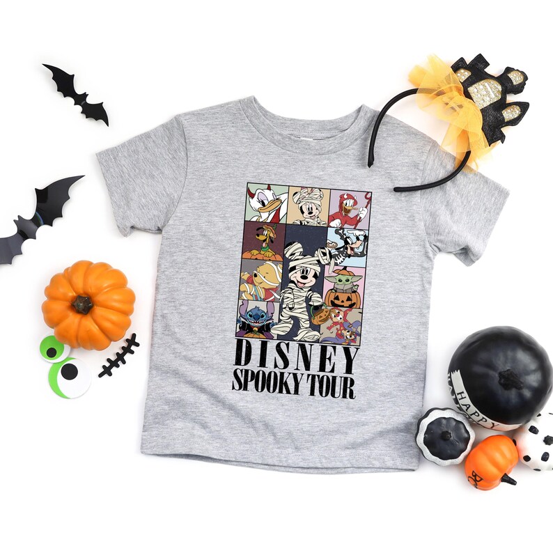 Pu&ograve; includere: Una maglietta grigia con una grafica colorata di personaggi Disney in costume di Halloween. La maglietta ha la scritta "DISNEY SPOOKY TOUR" in nero. Decorazioni di Halloween, tra cui zucche e pipistrelli, circondano la maglietta.