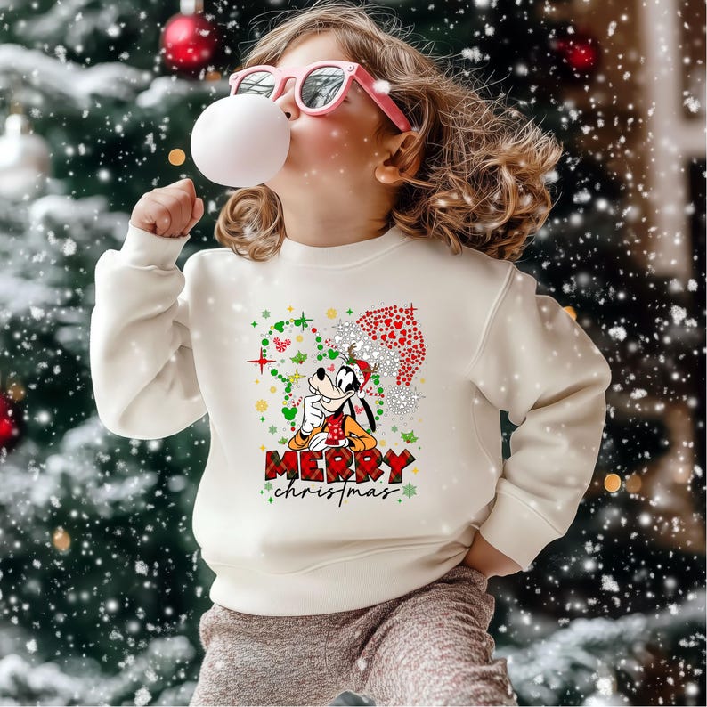 May include: A cream-colored sweatshirt featuring a graphic of Goofy in a Santa hat with the words "Merry Christmas." The design includes snowflakes and a red and green plaid pattern. The background is a snowy Christmas tree.