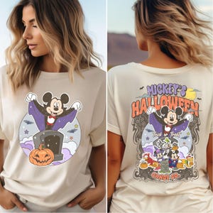 May include: A beige t-shirt featuring a vintage-style graphic of Mickey Mouse dressed as a vampire. The graphic says "Mickey's Halloween" and "Trick or Treat" with a tombstone that says "Candy Co."