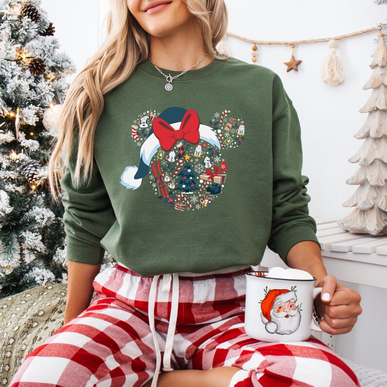 May include: Green sweatshirt with a festive Mickey Mouse head design, complete with a Santa hat and bow. The design incorporates Christmas elements like trees and snowflakes. The person is wearing red and white plaid pajama bottoms and holding a mug with a Santa Claus graphic.