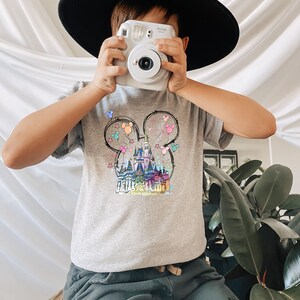 Op de afbeelding: Een grijs T-shirt met een kleurrijke illustratie van Assepoester's kasteel in de vorm van Mickey Mouse oren. Het kasteel is omringd door ballonnen en vuurwerk.