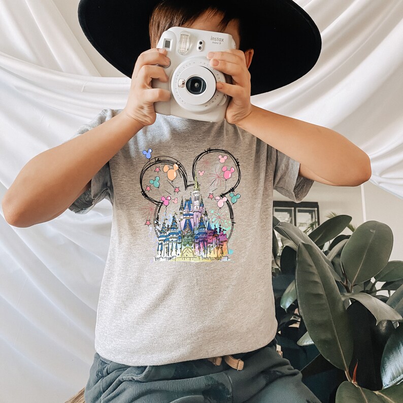Op de afbeelding: Een grijs T-shirt met een kleurrijke illustratie van Assepoester's kasteel in de vorm van Mickey Mouse oren. Het kasteel is omringd door ballonnen en vuurwerk.