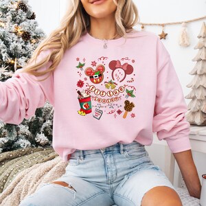 May include: A light pink sweatshirt featuring a festive graphic with the text "'Tis the Season". The design includes Mickey Mouse ears, a hot chocolate, and other holiday-themed illustrations. The sweatshirt is a long-sleeved crew neck.