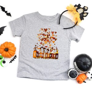 May include: A gray t-shirt featuring a Thanksgiving-themed graphic. The design includes cartoon characters, heart-shaped balloons in fall colors, and the word "Thanksgiving" in orange and brown. Decorative pumpkins and Halloween accessories surround the shirt.