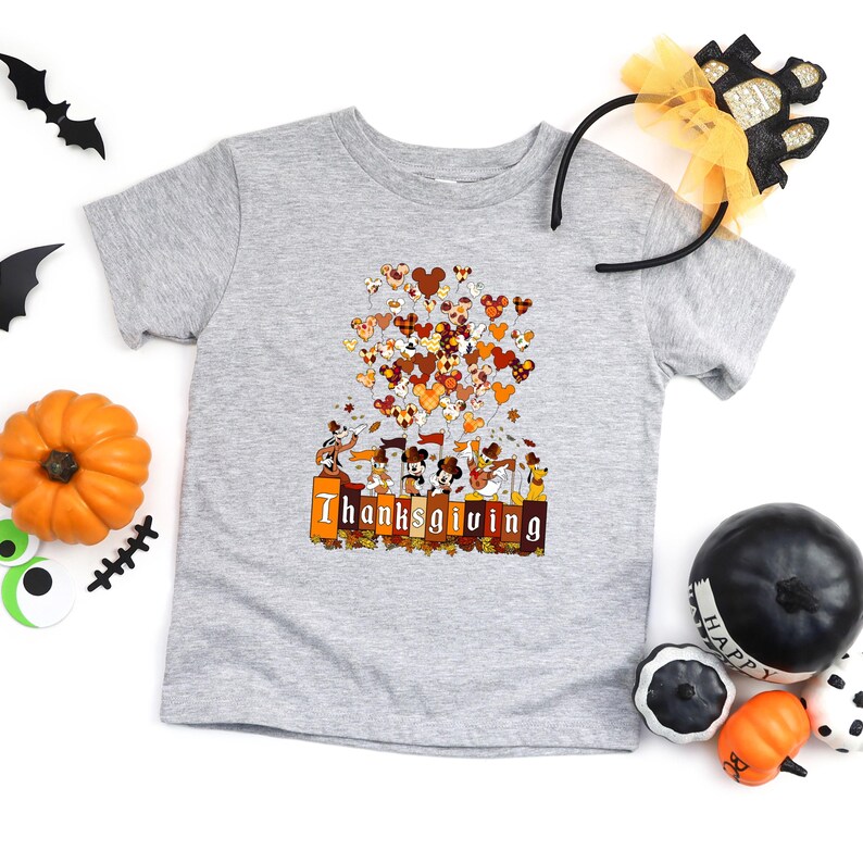 May include: A gray t-shirt featuring a Thanksgiving-themed graphic. The design includes cartoon characters, heart-shaped balloons in fall colors, and the word "Thanksgiving" in orange and brown. Decorative pumpkins and Halloween accessories surround the shirt.