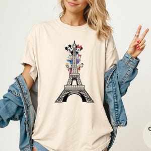 May include: Ivory-colored t-shirt featuring a black graphic of the Eiffel Tower with Disney characters. The shirt has the words "Comfort Colors Ivory" printed on the bottom right. The model is wearing a denim jacket.