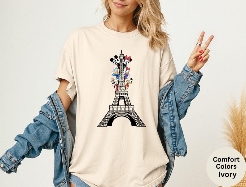 May include: Ivory-colored t-shirt featuring a black graphic of the Eiffel Tower with Disney characters. The shirt has the words "Comfort Colors Ivory" printed on the bottom right. The model is wearing a denim jacket.