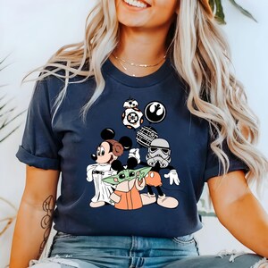 May include: Navy blue t-shirt featuring a graphic of Mickey Mouse, Baby Yoda, and a Stormtrooper. The design includes Star Wars and Disney elements. The shirt has a crew neck and short sleeves.