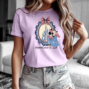 May include: A light purple t-shirt with a graphic of Mickey Mouse in front of Cinderella's castle. The graphic is framed with a blue and gold ornate frame and a pink bow. The text "Happiest place on earth" is printed below the graphic.