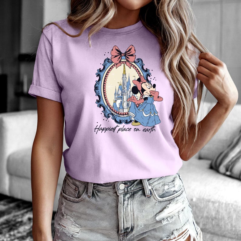 May include: A light purple t-shirt with a graphic of Mickey Mouse in front of Cinderella's castle. The graphic is framed with a blue and gold ornate frame and a pink bow. The text "Happiest place on earth" is printed below the graphic.