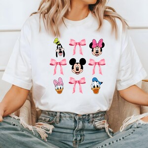 May include: White t-shirt with a graphic print of Mickey Mouse, Minnie Mouse, Donald Duck, Goofy, and Daisy Duck. Each character is surrounded by a pink bow.