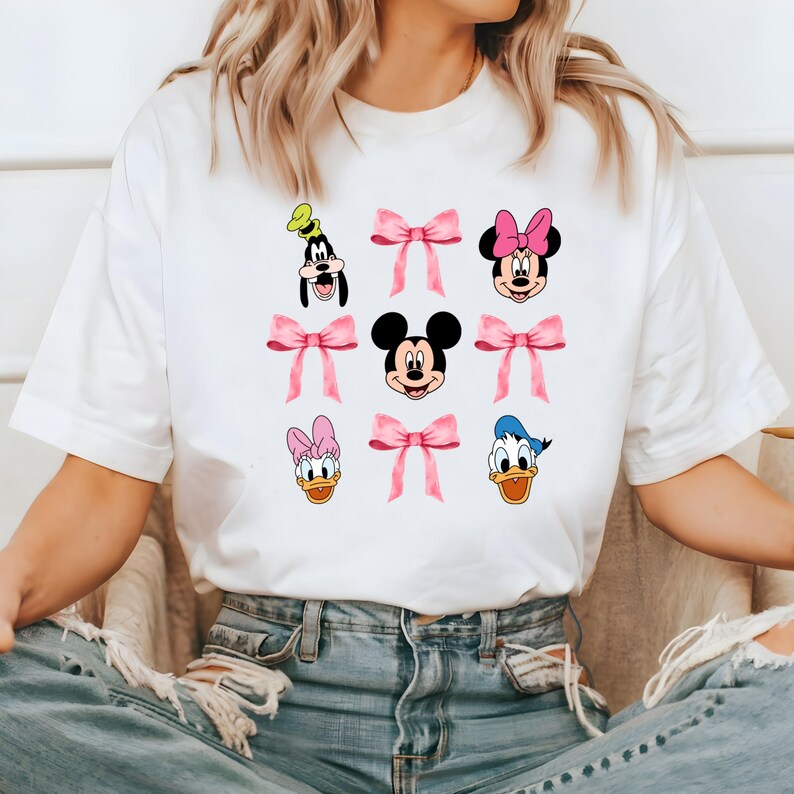 May include: White t-shirt with a graphic print of Mickey Mouse, Minnie Mouse, Donald Duck, Goofy, and Daisy Duck. Each character is surrounded by a pink bow.