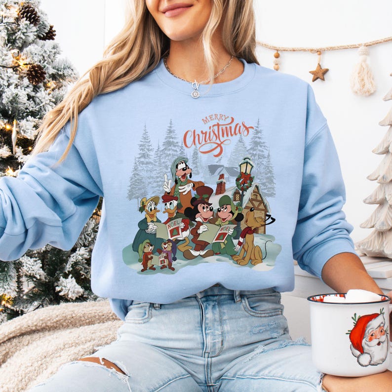 May include: Light blue sweatshirt featuring a festive Christmas scene with cartoon characters. The graphic includes Mickey Mouse, Donald Duck, Goofy, and others, gathered in a snowy landscape. The words "Merry Christmas" are written in red script above the scene.