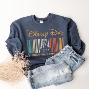 May include: A navy blue sweatshirt with the text "Disney Dad" and a vintage-style barcode with a silhouette of Cinderella's castle and Mickey Mouse. The text "Scan for Payment" is below the barcode.