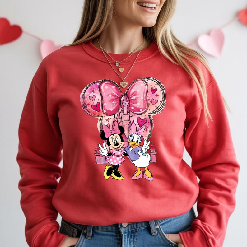 May include: Red sweatshirt featuring a Minnie Mouse and Daisy Duck graphic. The graphic includes a pink bow, hearts, and a castle design. Minnie Mouse is wearing a pink polka dot dress, and Daisy Duck is wearing a purple top and pink shoes.