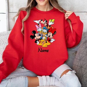 Op de afbeelding: Rood sweatshirt met een afbeelding van Mickey Mouse, Minnie Mouse, Donald Duck, Goofy en Pluto. De tekst "NAME" is onder de afbeelding gedrukt.