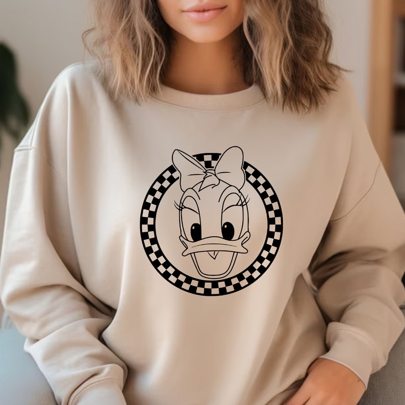 May include: A beige sweatshirt with a black and white chequered circle design featuring a cartoon duck with a bow.