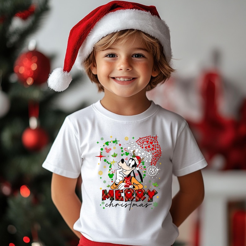 May include: A child wearing a white t-shirt with a Christmas graphic featuring a cartoon dog in a Santa hat and the words "Merry Christmas". The child is also wearing a red Santa hat.