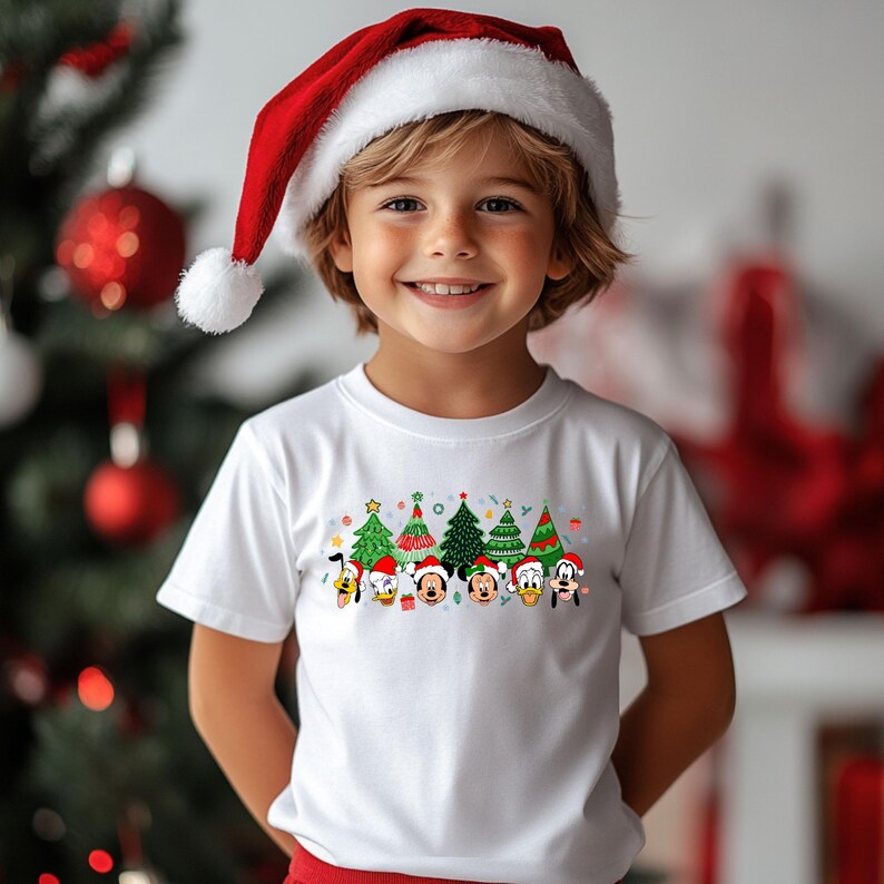 May include: A young child wearing a white t-shirt with a Christmas tree design and a Santa hat. The shirt features cartoon characters with Christmas tree designs. The child is smiling in front of a Christmas tree.