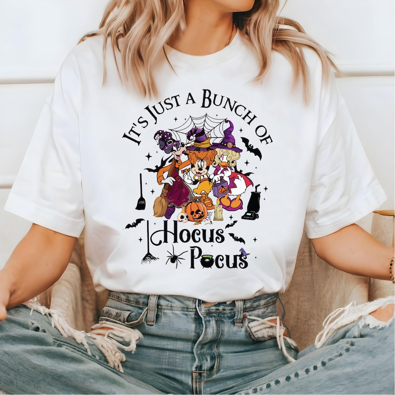 May include: White t-shirt with a graphic of Mickey Mouse, Minnie Mouse, and Donald Duck dressed as witches. The text "It's Just a Bunch of Hocus Pocus" is printed on the shirt.