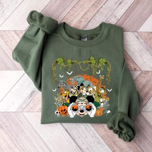 May include: Olive green sweatshirt featuring a safari-themed graphic. The design includes cartoon animals like a zebra, elephant, and giraffe, along with Mickey Mouse and friends. The graphic also includes pumpkins, bats, and spiderwebs.