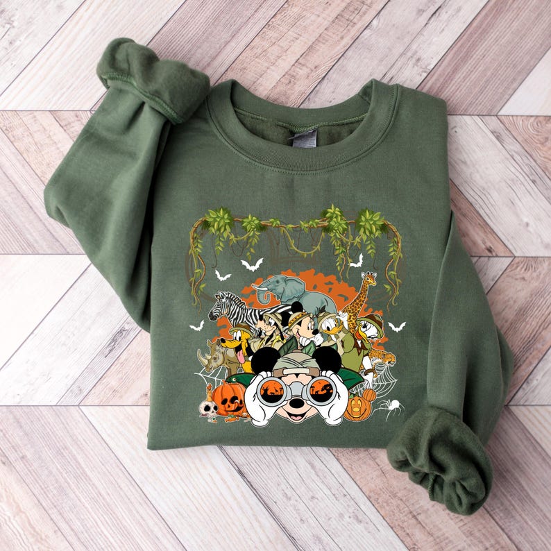 May include: Olive green sweatshirt featuring a safari-themed graphic. The design includes cartoon animals like a zebra, elephant, and giraffe, along with Mickey Mouse and friends. The graphic also includes pumpkins, bats, and spiderwebs.