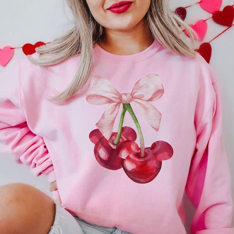 Puede incluir: Sudadera rosa con un gr&aacute;fico de dos cerezas rojas con un lazo rosa en la parte superior.