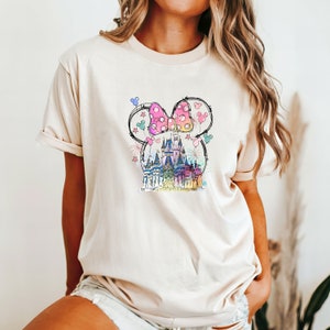 Op de afbeelding: Een beige T-shirt met een kleurrijke afbeelding van een kasteel, een strik en ballonnen. De afbeelding is in cartoon-stijl en heeft een roze strik met witte stippen.