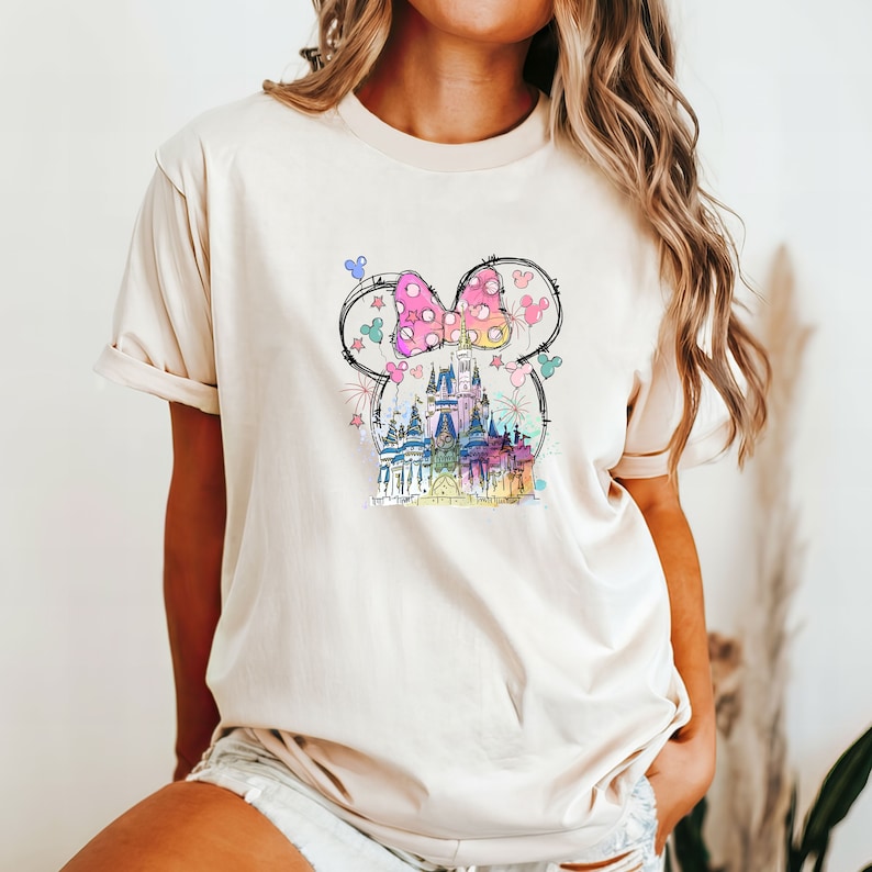 Op de afbeelding: Een beige T-shirt met een kleurrijke afbeelding van een kasteel, een strik en ballonnen. De afbeelding is in cartoon-stijl en heeft een roze strik met witte stippen.