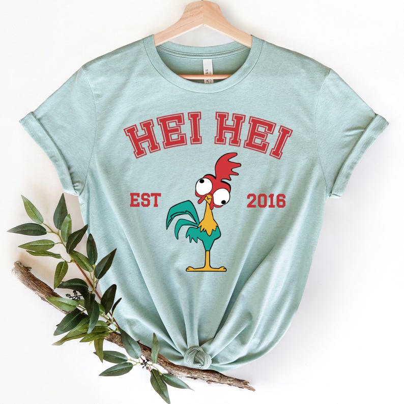 以下が含まれることがあります： 薄い青色のTシャツに、赤い鶏のグラフィックと「Hei Hei Est 2016」の文字がプリントされています。