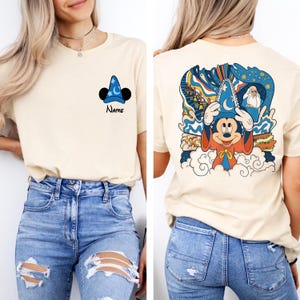 May include: A light beige t-shirt with a cartoon image of Mickey Mouse wearing a sorcerer's hat on the front. The back of the shirt features a colorful illustration of Mickey Mouse as a sorcerer with a whimsical background.
