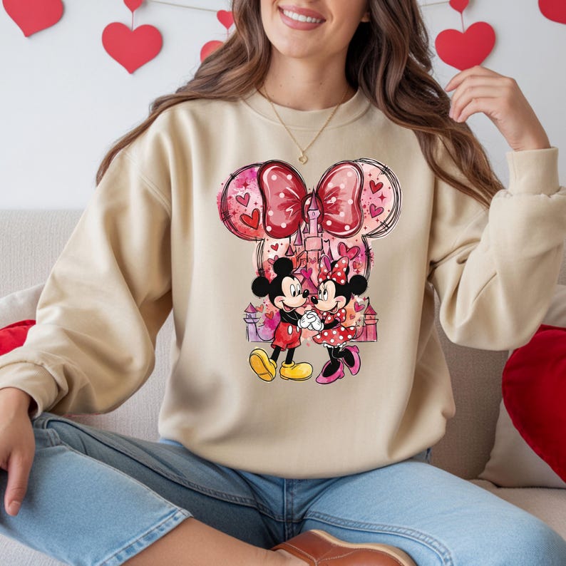 May include: Beige sweatshirt featuring a colorful graphic of Mickey and Minnie Mouse holding hands in front of a castle. The design includes a large bow and hearts. The sweatshirt is a long-sleeved, crew neck style.