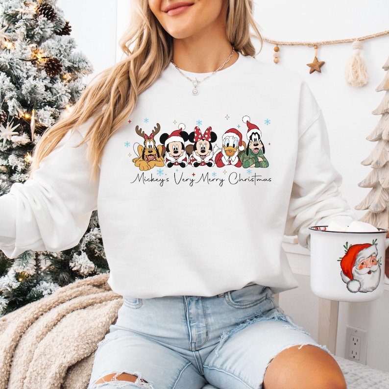 Puede incluir: Sudadera blanca con un dise&ntilde;o festivo de personajes de Disney con atuendos navide&ntilde;os. El dise&ntilde;o incluye a Mickey, Minnie, Donald, Goofy y Pluto, con el texto "Mickey's Very Merry Christmas". Tambi&eacute;n se ve una taza tem&aacute;tica navide&ntilde;a.