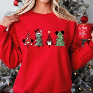 May include: Red Christmas sweatshirt with a graphic of five Christmas trees decorated with Mickey Mouse ears and ornaments. The trees are in a row and the text "merry + bright" is on a mug in the background.
