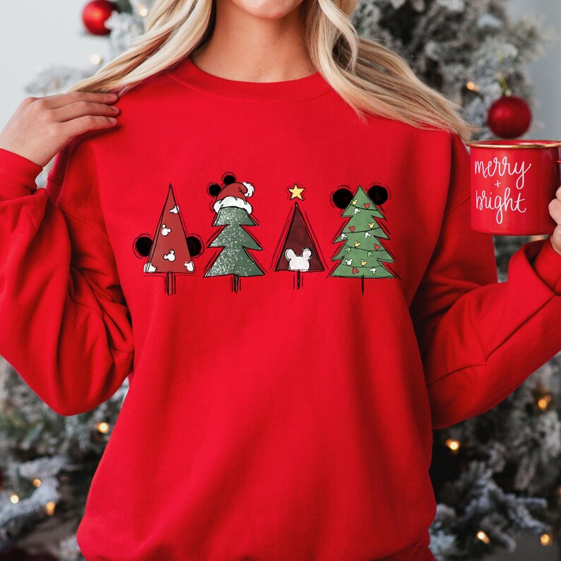 May include: Red Christmas sweatshirt with a graphic of five Christmas trees decorated with Mickey Mouse ears and ornaments. The trees are in a row and the text "merry + bright" is on a mug in the background.
