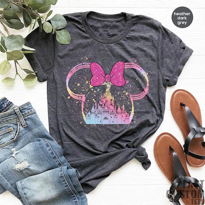 May include: A heather dark grey t-shirt with a pink bow and a colorful castle graphic inside a Mickey Mouse ear silhouette.