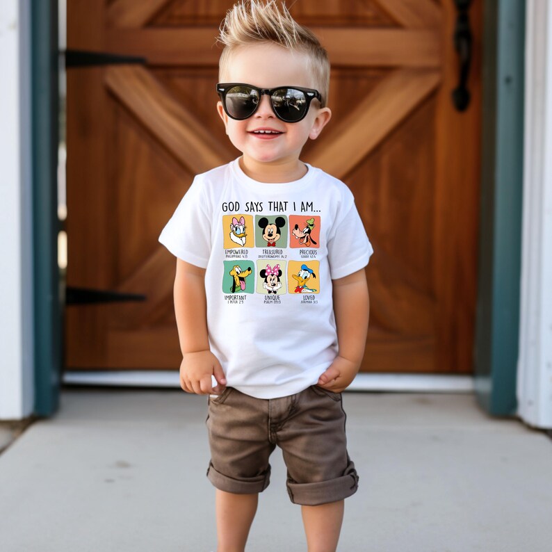 Puede incluir: Una camiseta blanca con personajes de Disney y el texto "GOD SAYS THAT I AM...". La camiseta presenta a Daisy Duck, Mickey Mouse y Goofy en la parte superior, y a Pluto, Minnie Mouse y Donald Duck en la parte inferior. El ni&ntilde;o lleva pantalones cortos marrones y gafas de sol.