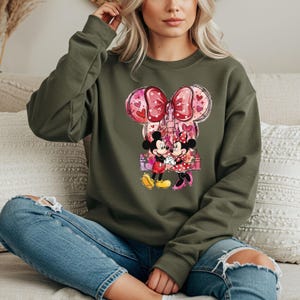 May include: An olive green sweatshirt featuring a graphic of Mickey and Minnie Mouse holding hands in front of a castle. The design includes a large pink bow with hearts and a castle silhouette. The characters are in their classic outfits.