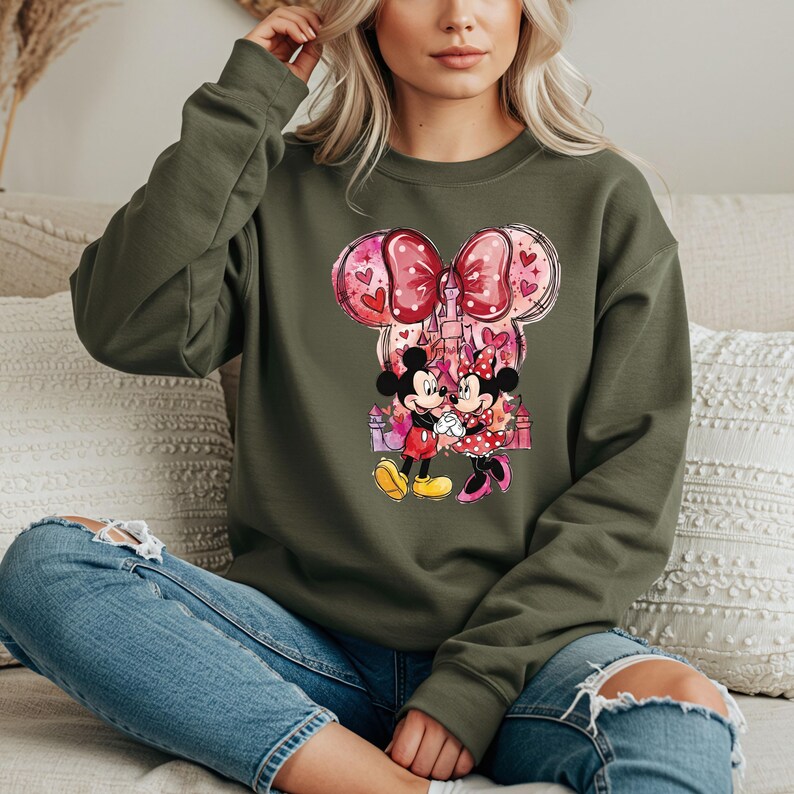 May include: An olive green sweatshirt featuring a graphic of Mickey and Minnie Mouse holding hands in front of a castle. The design includes a large pink bow with hearts and a castle silhouette. The characters are in their classic outfits.