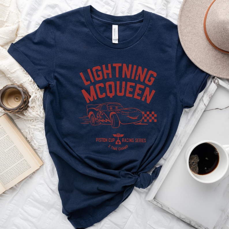 May include: A navy blue t-shirt with a red graphic of Lightning McQueen from the Disney Pixar Cars movie. The graphic includes the text "Lightning McQueen", "Piston Cup", "Racing Series", and "5 Time Champ".