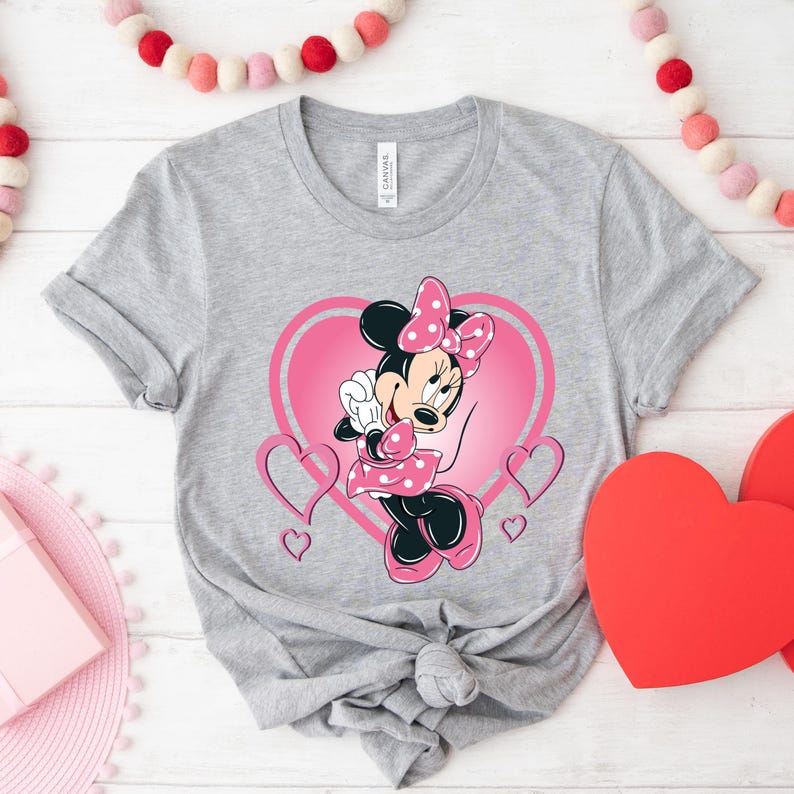 May include: A gray t-shirt with a pink heart and Minnie Mouse graphic. Minnie Mouse is wearing a pink dress with white polka dots and a pink bow.