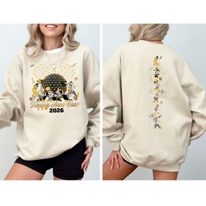 May include: Beige sweatshirt with a graphic featuring Disney characters, a golden sphere, and the text "Happy New Year 2026." The back of the sweatshirt has a vertical line of Disney character illustrations.