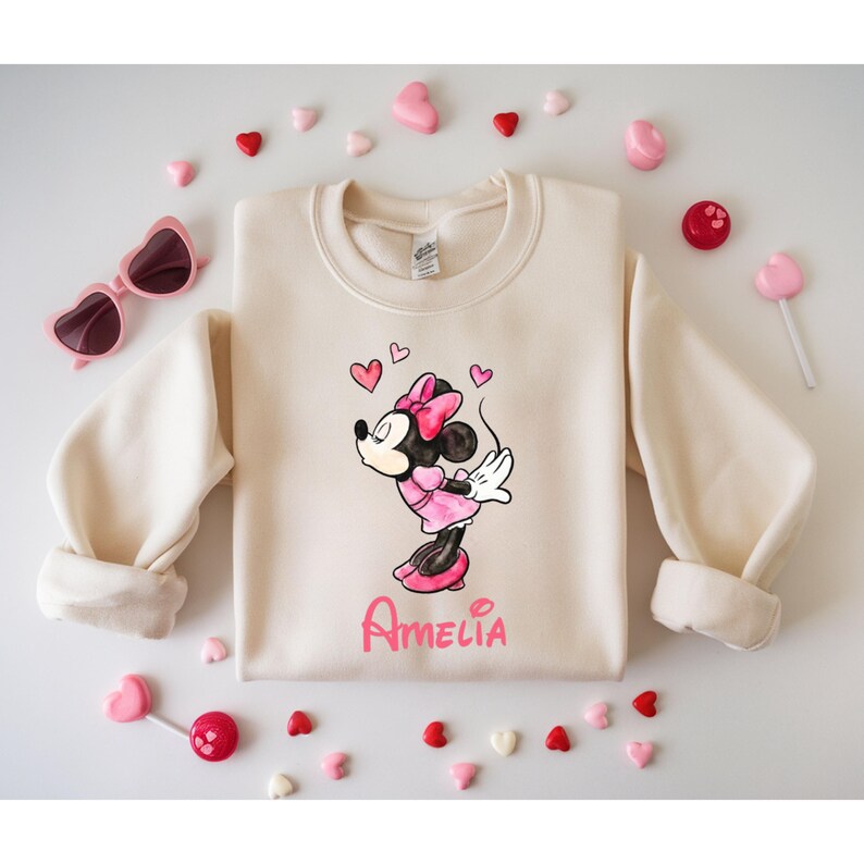 May include: Cream-colored sweatshirt featuring a Minnie Mouse graphic in a pink dress and bow, with the name "Amelia" in pink script. Pink heart-shaped candies and sunglasses surround the sweatshirt, along with heart-shaped lollipops.