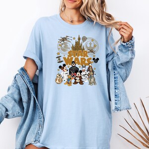May include: Light blue t-shirt featuring a Star Wars graphic with Disney characters. The design includes the Star Wars logo, a castle, spaceships, and characters like Mickey Mouse, Darth Vader, and R2-D2. The shirt has short sleeves.