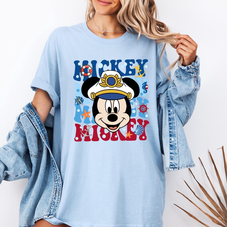 May include: Light blue t-shirt with a cartoon image of Mickey Mouse wearing a captain's hat and surrounded by nautical themed graphics. The text "Mickey Mouse" is printed in red, white, and blue letters.