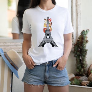 May include: White t-shirt featuring a black Eiffel Tower design with cartoon princess characters. The shirt is a crew neck and has short sleeves. The model is wearing denim shorts.