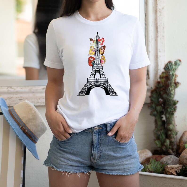May include: White t-shirt featuring a black Eiffel Tower design with cartoon princess characters. The shirt is a crew neck and has short sleeves. The model is wearing denim shorts.