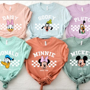 May include: A collection of six colorful t-shirts featuring Disney characters: Daisy, Goofy, Pluto, Donald, Minnie, and Mickey. Each shirt displays the character's name, face, and the year they debuted, set against a checkered background. The shirts are in various pastel colors.
