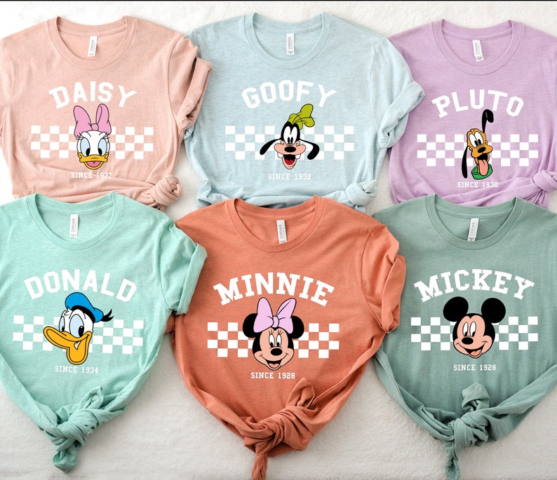 May include: A collection of six colorful t-shirts featuring Disney characters: Daisy, Goofy, Pluto, Donald, Minnie, and Mickey. Each shirt displays the character's name, face, and the year they debuted, set against a checkered background. The shirts are in various pastel colors.
